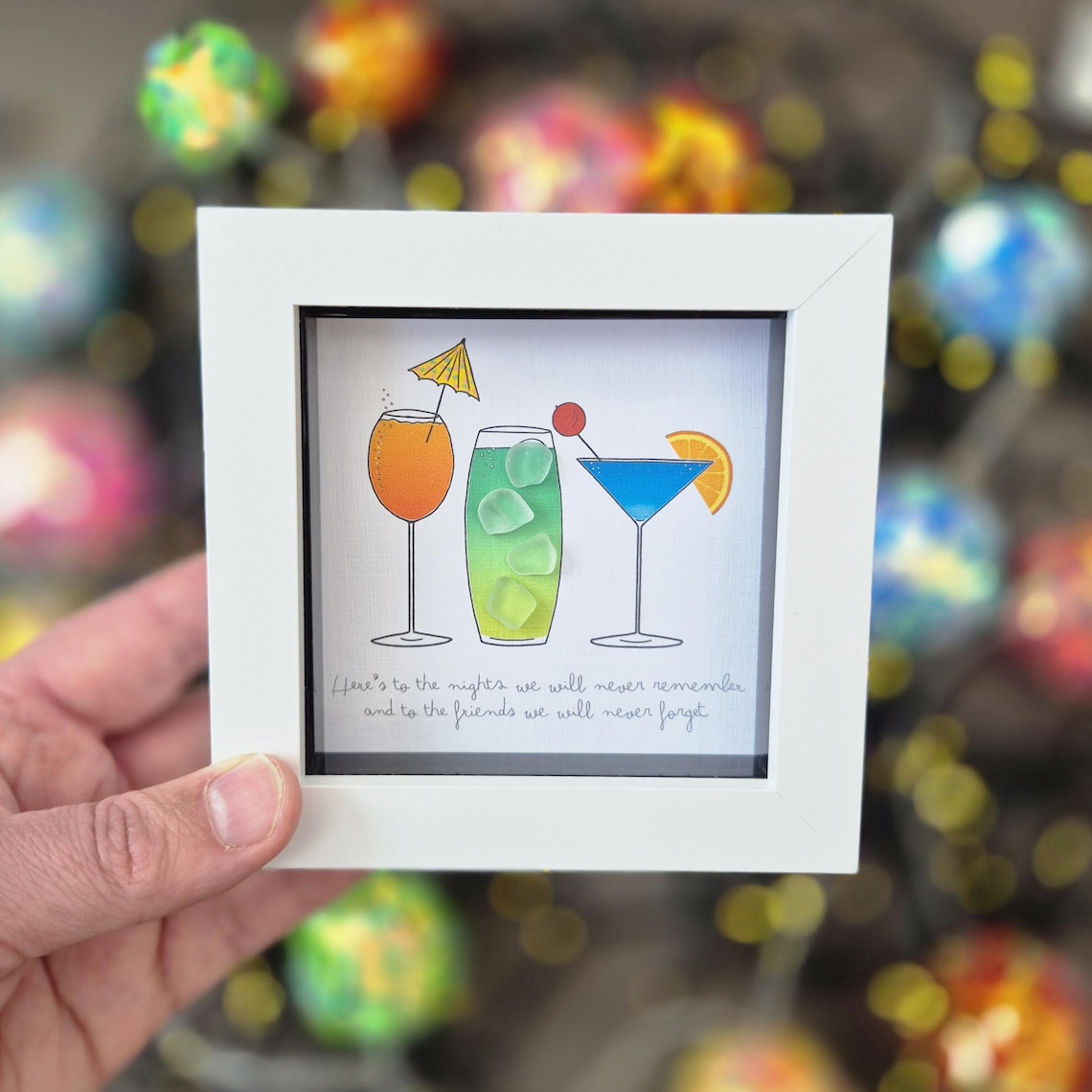 May include: White framed art print featuring three colorful cocktail illustrations. The drinks include an orange cocktail with a yellow umbrella, a green cocktail with ice, and a blue cocktail with an orange slice. The text reads, "Here's to the nights we will never remember and to the friends we will never forget."