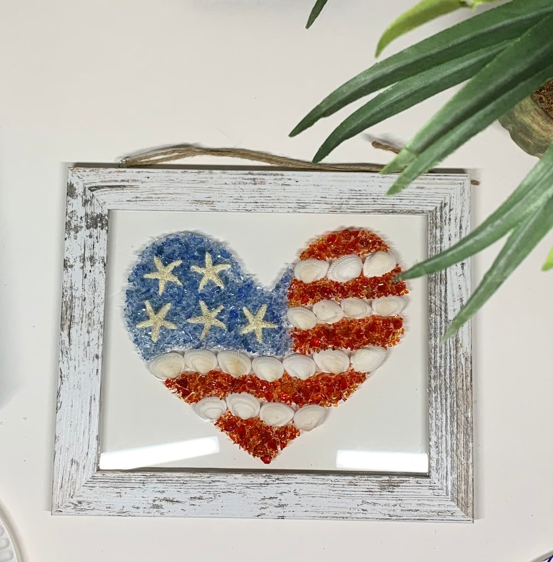 May include: A white wooden frame with a glass front, showcasing a heart-shaped American flag made of seashells and glass beads. The flag is red, white, and blue, with the stars made of white seashells and the stripes made of red and white seashells.