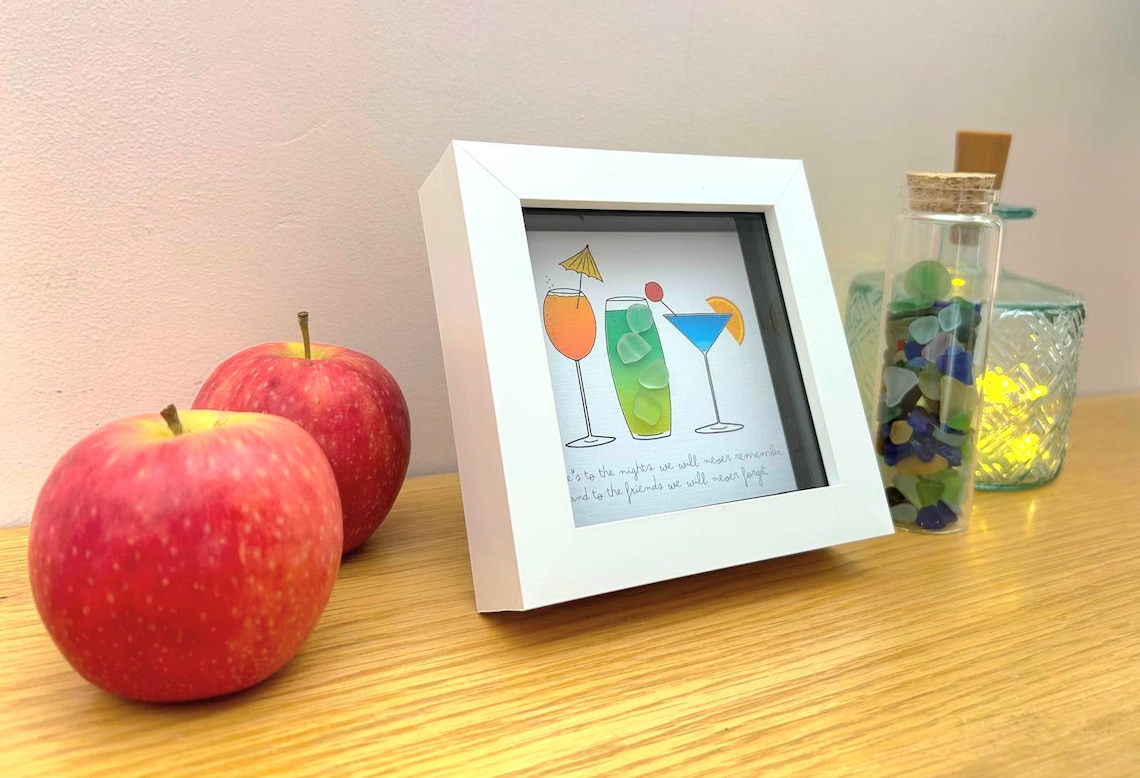 May include: A white framed print with cocktail illustrations, two red apples, and a glass bottle filled with colorful stones. The print has text at the bottom. A decorative glass jar is also visible.