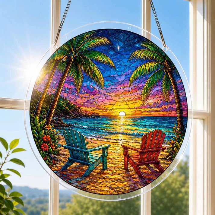 A round acrylic suncatcher with a tropical beach sunset two adirondack chairs and palm trees hanging from a chain in front of a bright white window