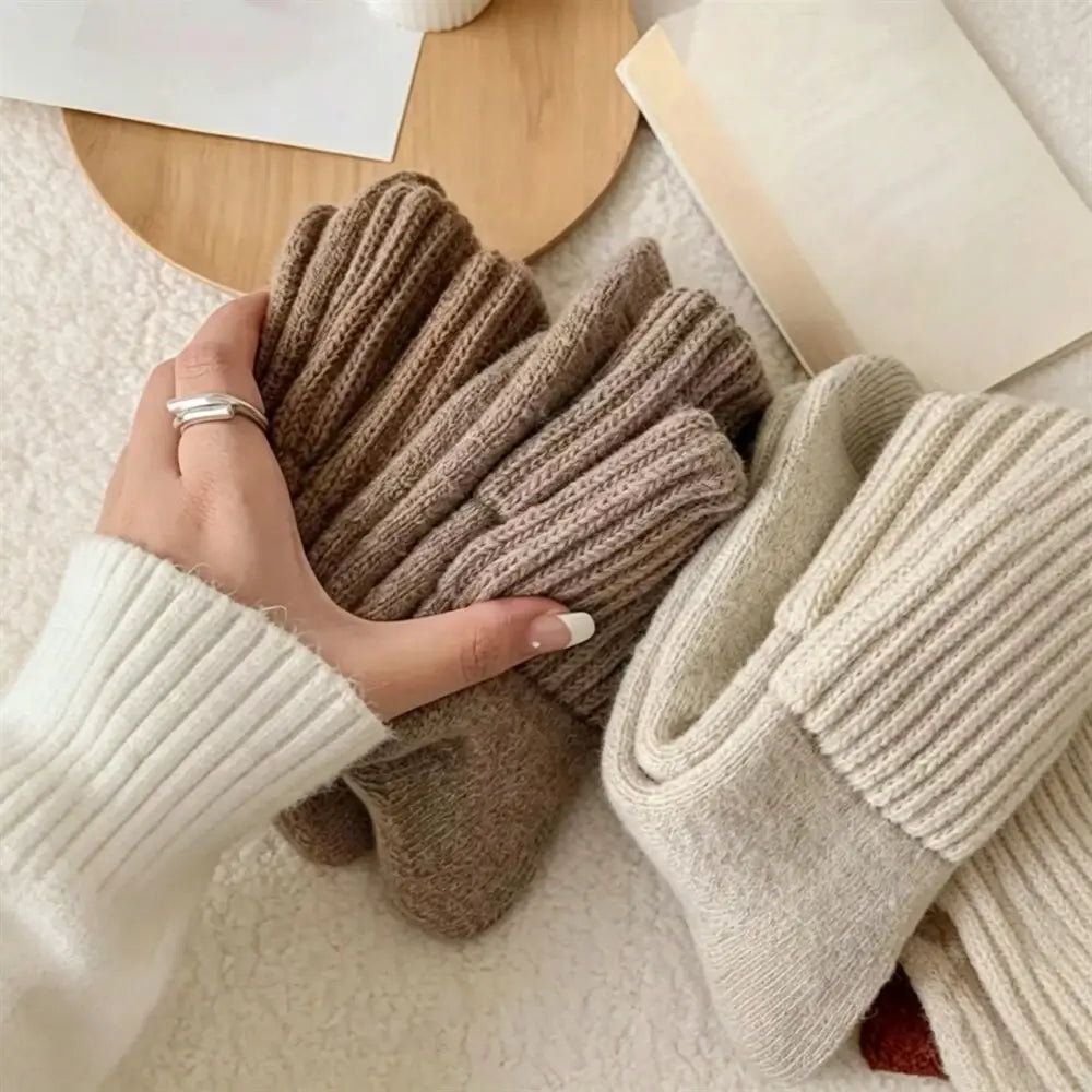 A hand wearing a silver ring holds a pair of ribbed knit gloves in taupe-brown, with two more pairs in cream and taupe resting on a soft beige surface. The scene includes a wooden tray and a book as styled props.