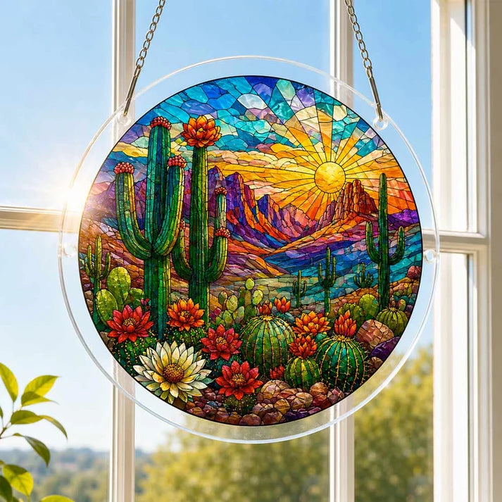 A round acrylic suncatcher with saguaro cacti blooming flowers and desert mountains hanging from a chain in front of a bright white window
