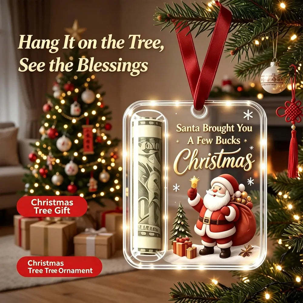 A Christmas tree ornament shown hanging in front of a decorated Christmas tree and wrapped gifts. The ornament is a clear rectangular plastic tag with a red ribbon loop, featuring a rolled dollar bill, Santa Claus, gifts, and the text 'Santa Brought You A Few Bucks Christmas.