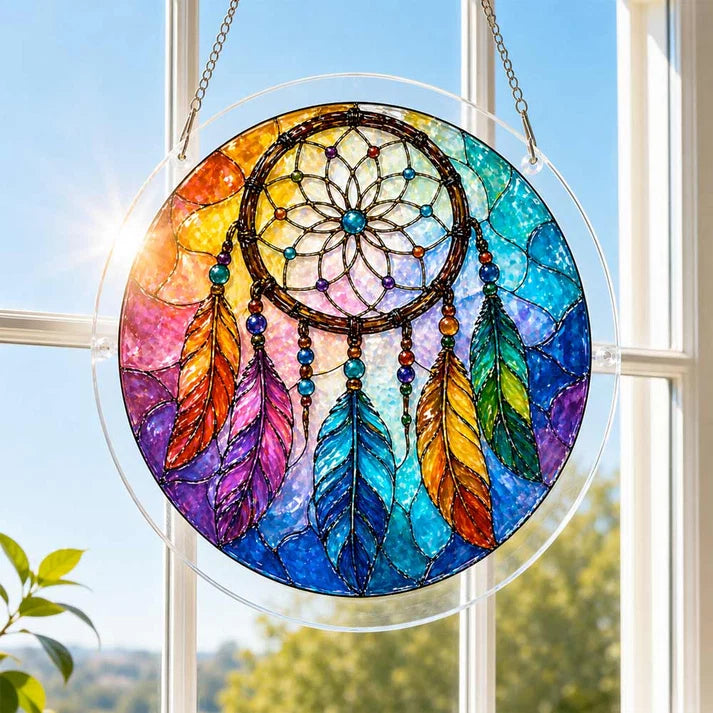 A round acrylic suncatcher with a beaded rainbow dreamcatcher and multicolored feathers hanging from a chain in front of a bright white window