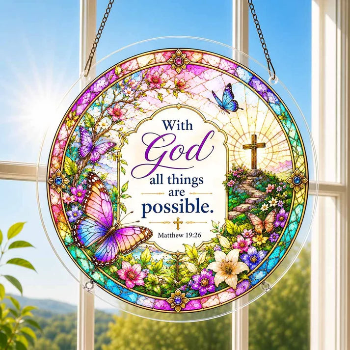 A round acrylic suncatcher featuring the verse with God all things are possible decorated with butterflies and flowers hanging from a chain in front of a bright white window