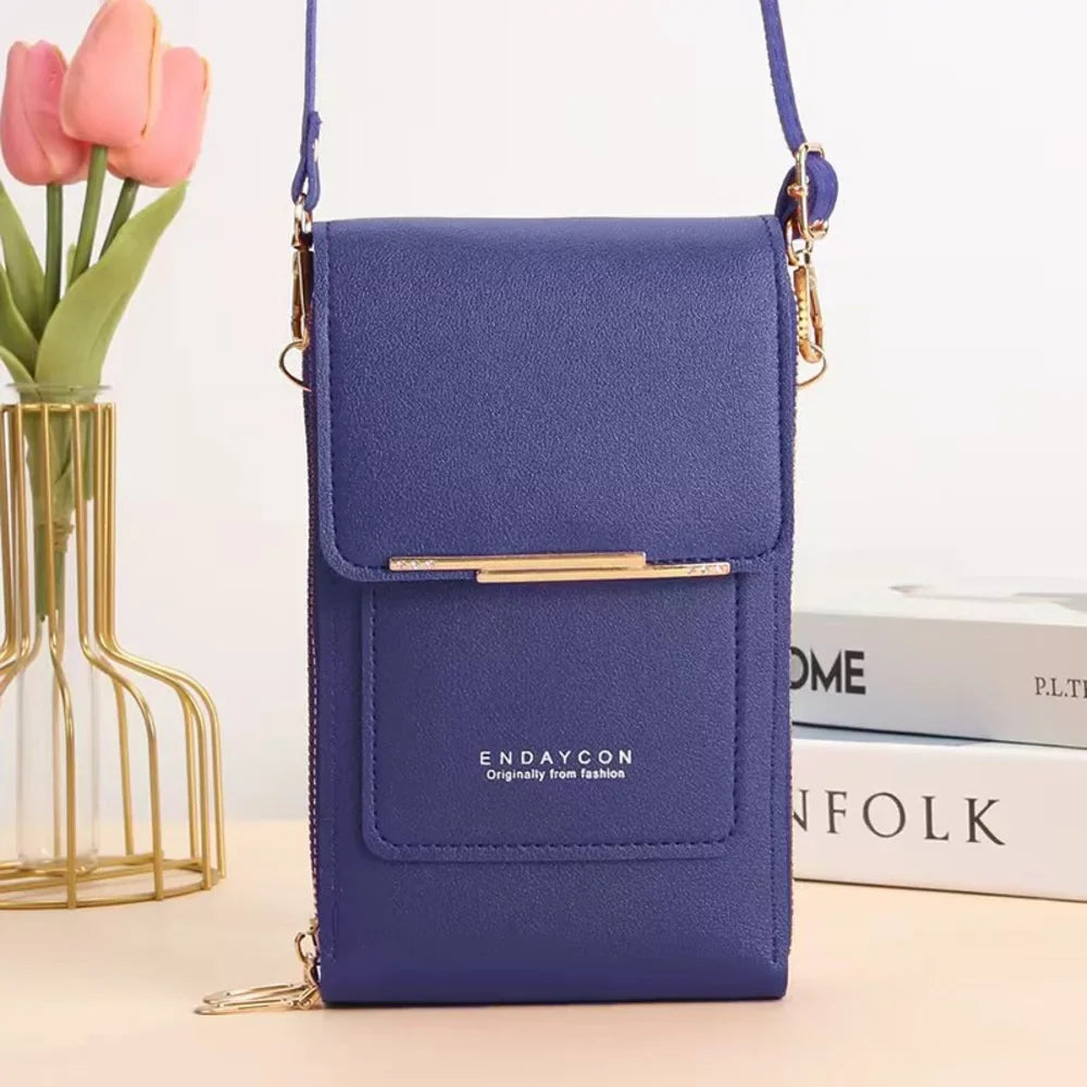 A royal blue leather crossbody phone bag with flap cover and front pocket beside a gold vase of pink tulips and white books