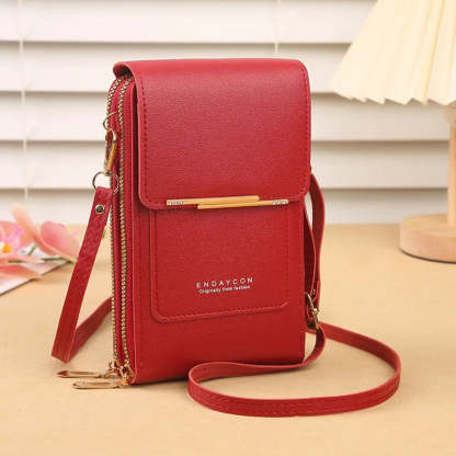 A red leather crossbody phone bag with flap cover rhinestone bar and double zippers on a beige desk with white blinds and pink flowers