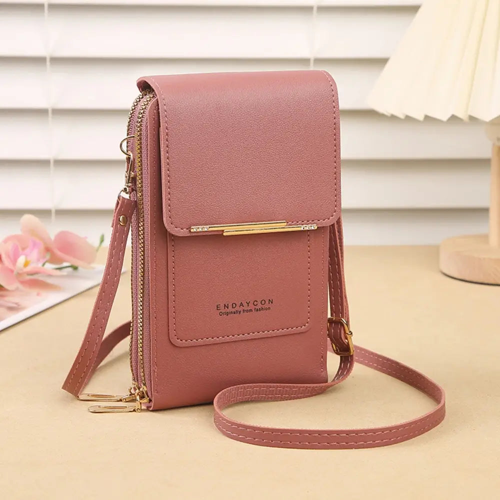 A rose pink leather crossbody phone bag with flap cover rhinestone bar and double zippers on a beige desk with white blinds and pink flowers