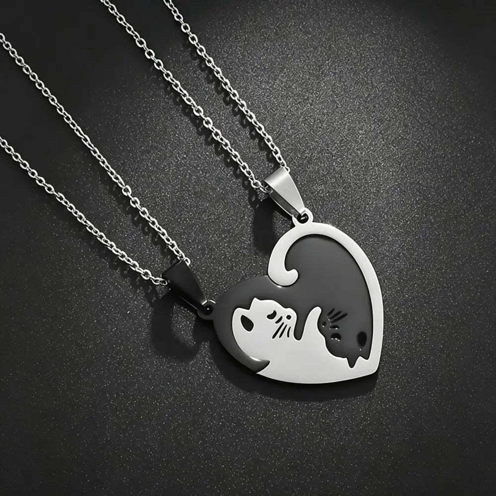 Two silver heart-shaped pendant necklaces on matching chains, designed to fit together into one heart. One pendant is polished silver with a black cat silhouette, and the other is black with a silver cat silhouette, set against a dark textured background.