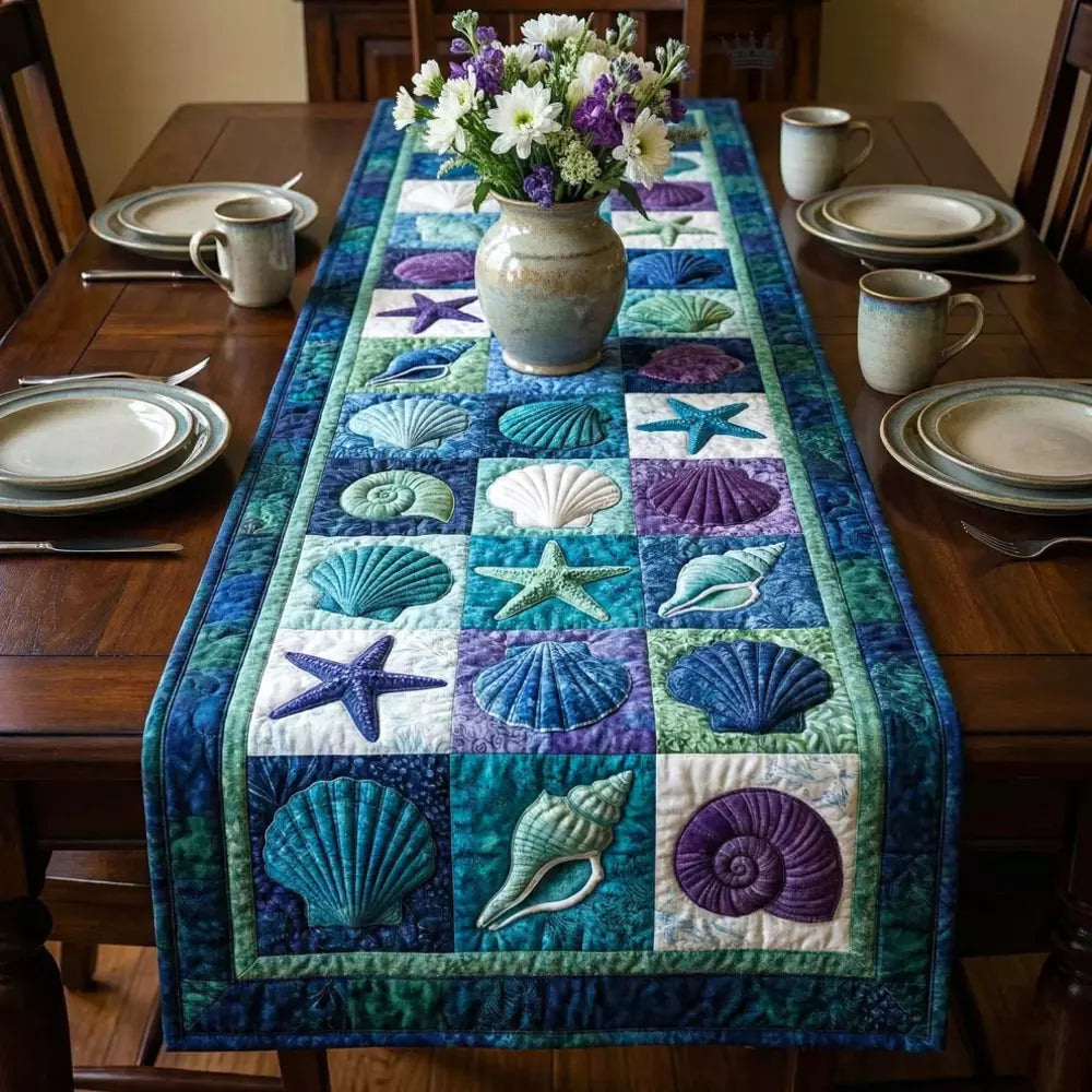 A quilted table runner with shells starfish and ocean patchwork squares laid on a wooden dining table with a ceramic vase of white daisies and purple flowers in the center