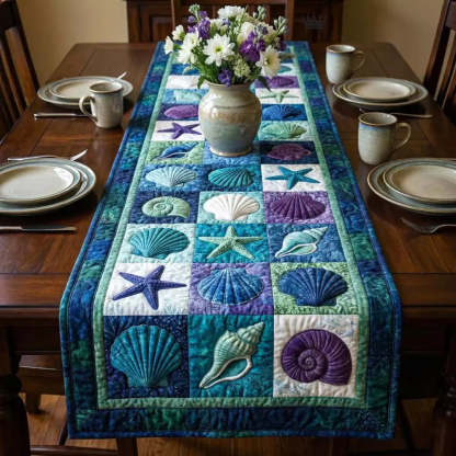 A quilted table runner with shells starfish and ocean patchwork squares laid on a wooden dining table with a ceramic vase of white daisies and purple flowers in the center