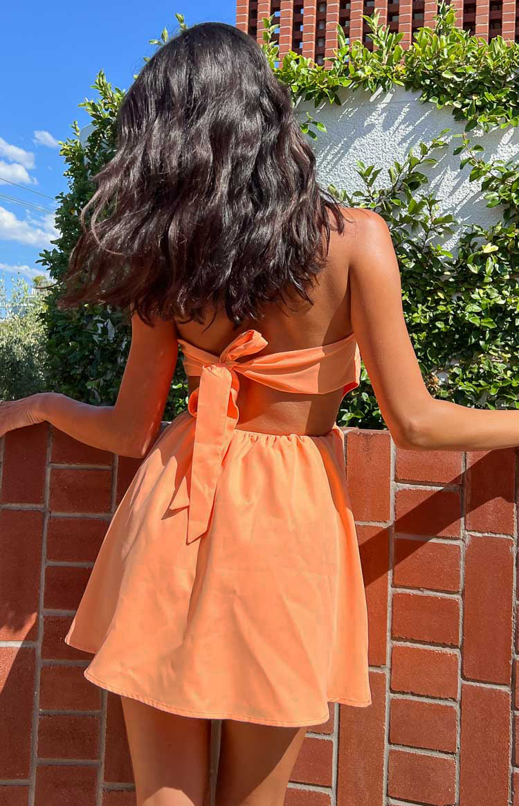 Model posing against white wall in orange strapless mini dress