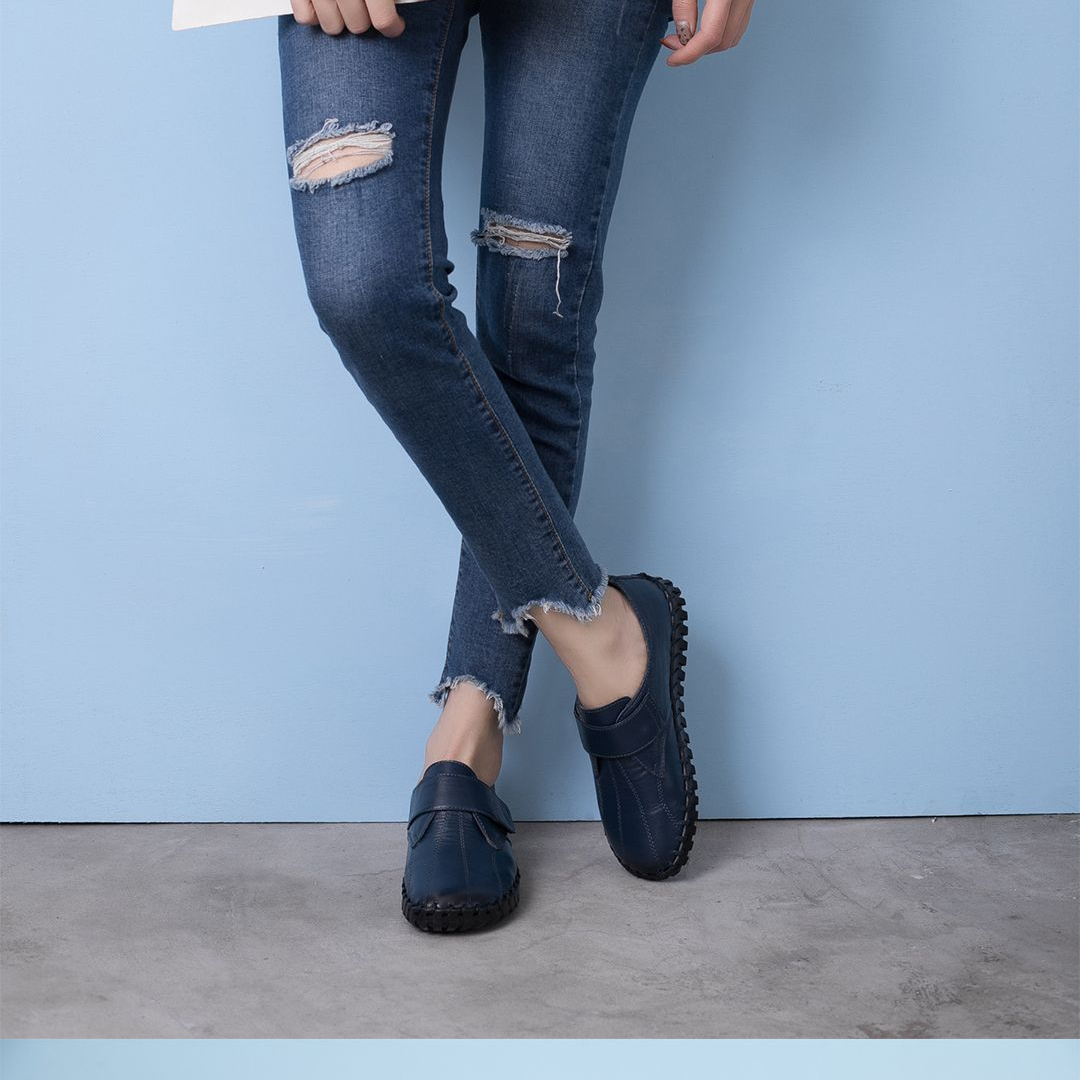 Piernas de una persona de pie con las piernas cruzadas que lleva vaqueros agujereados y zapatos azules.