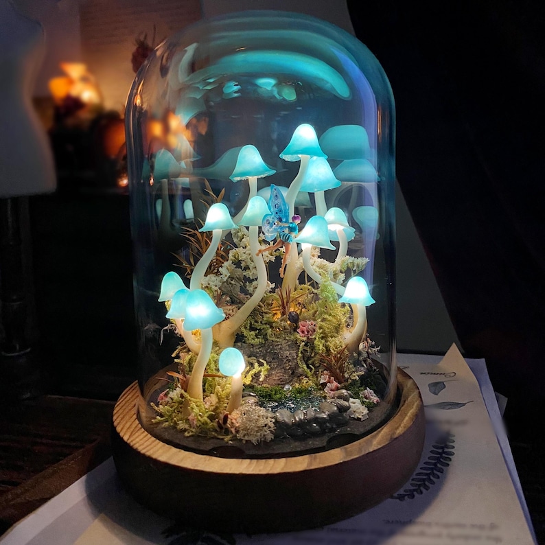 May include: A glass dome with a wooden base containing a miniature forest scene with glowing blue mushrooms and a blue fairy figurine.