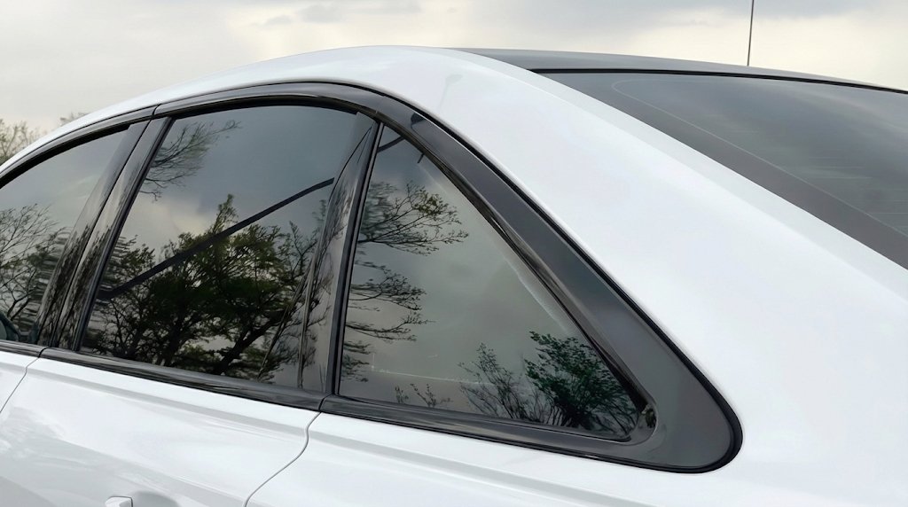 Close-up view of the rear side and C-pillar of a white sedan, featuring dark tinted windows with distinct tree reflections under a cloudy sky.