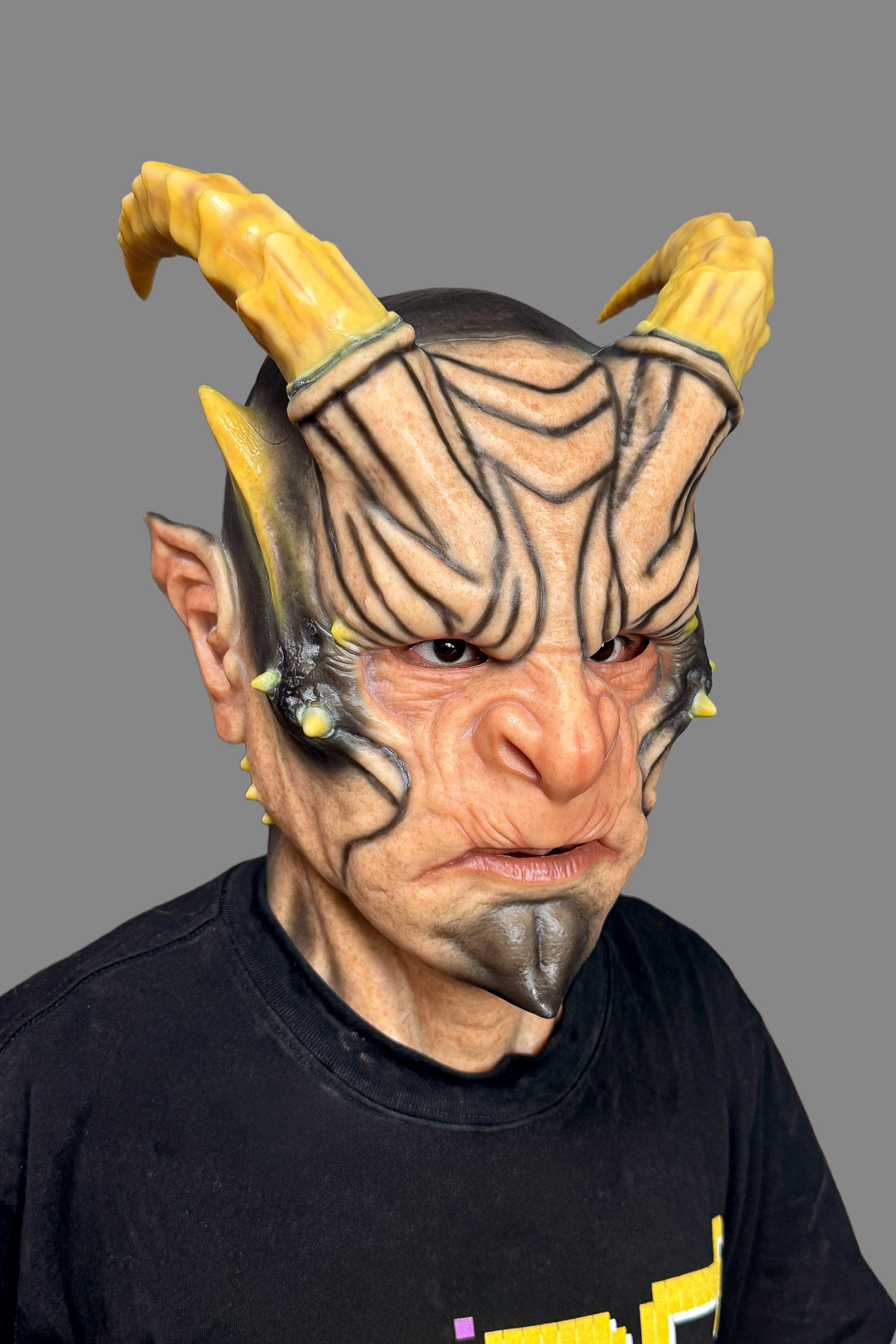 Realistic devil silicone full-head mask with curved yellow horns, pointed elf ears, tribal face markings and detailed skin texture, side view on model