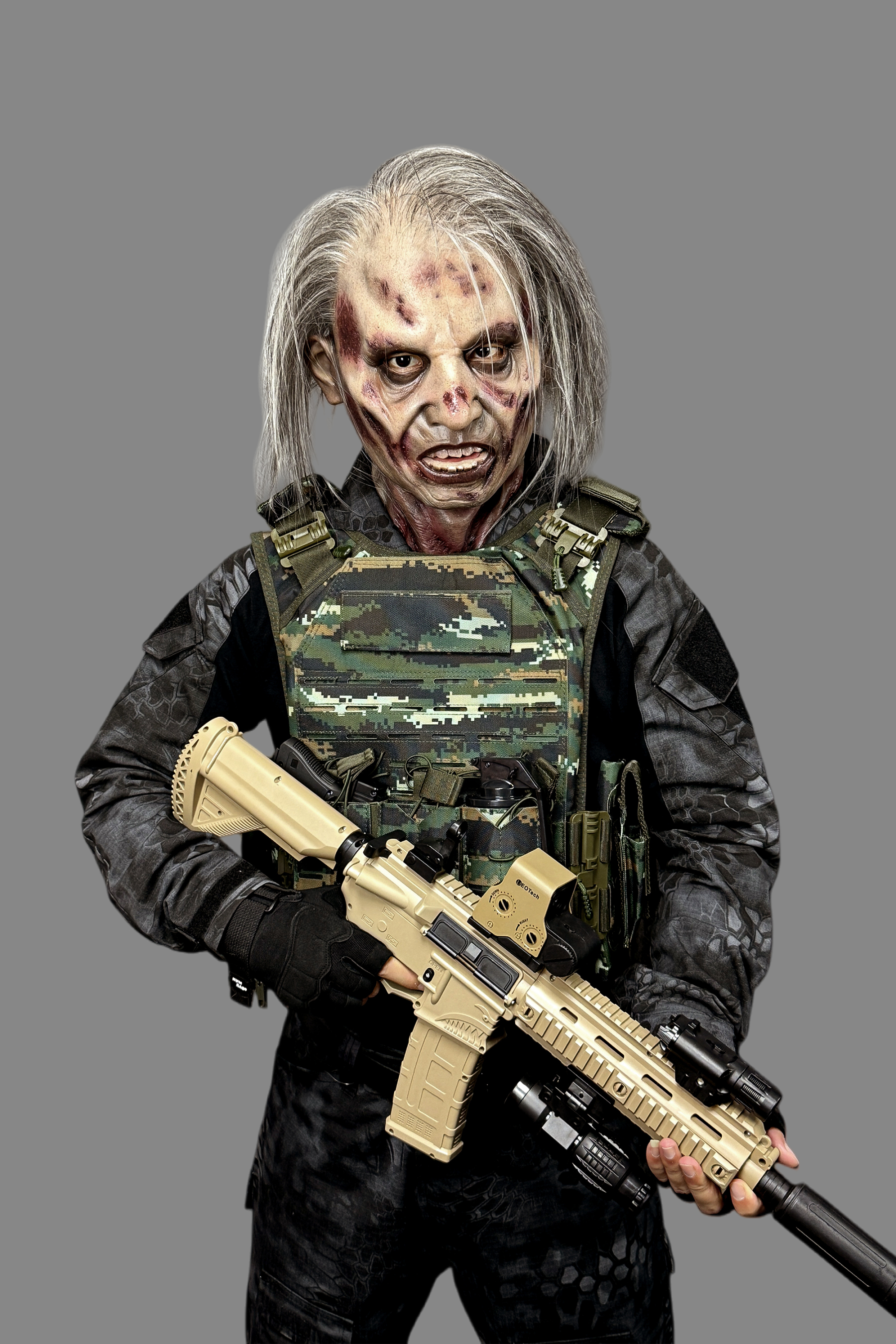 Scary zombie operator silicone mask with rotting flesh and long hair, posed with tan assault rifle, complete military tactical gear and plate carrier.