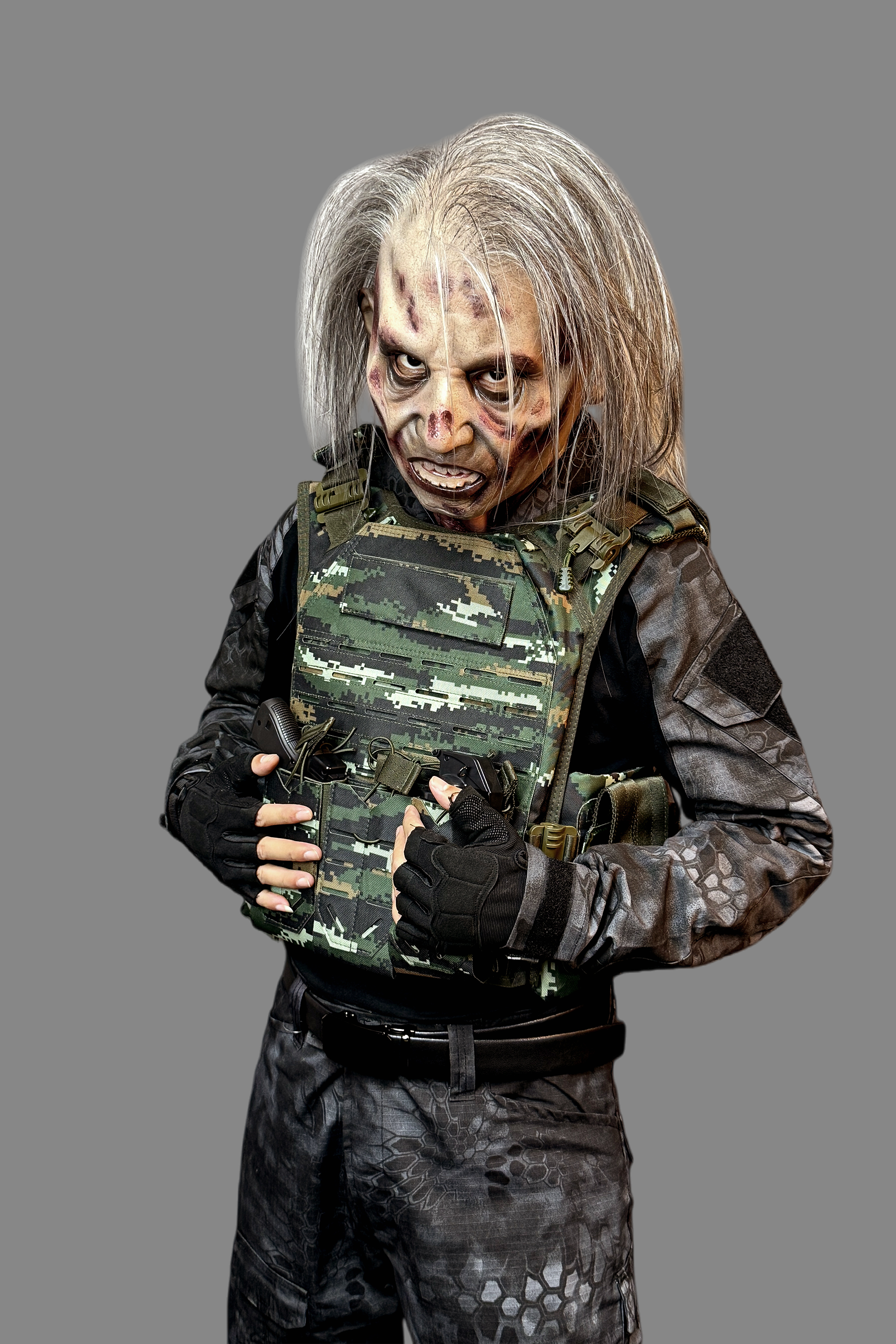 Lifelike undead elderly zombie silicone mask with long gray hair and realistic decay, worn in camouflage combat uniform, side angle.