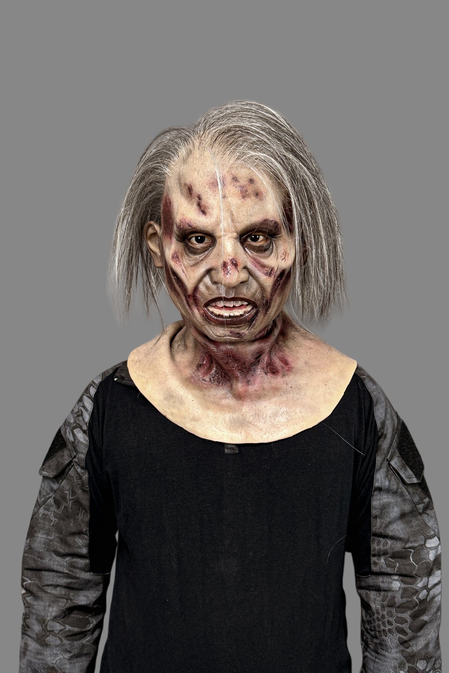 Hyper-realistic zombie old man silicone mask with long gray hair, rotting flesh, blood wounds and exposed teeth, worn with tactical black shirt.