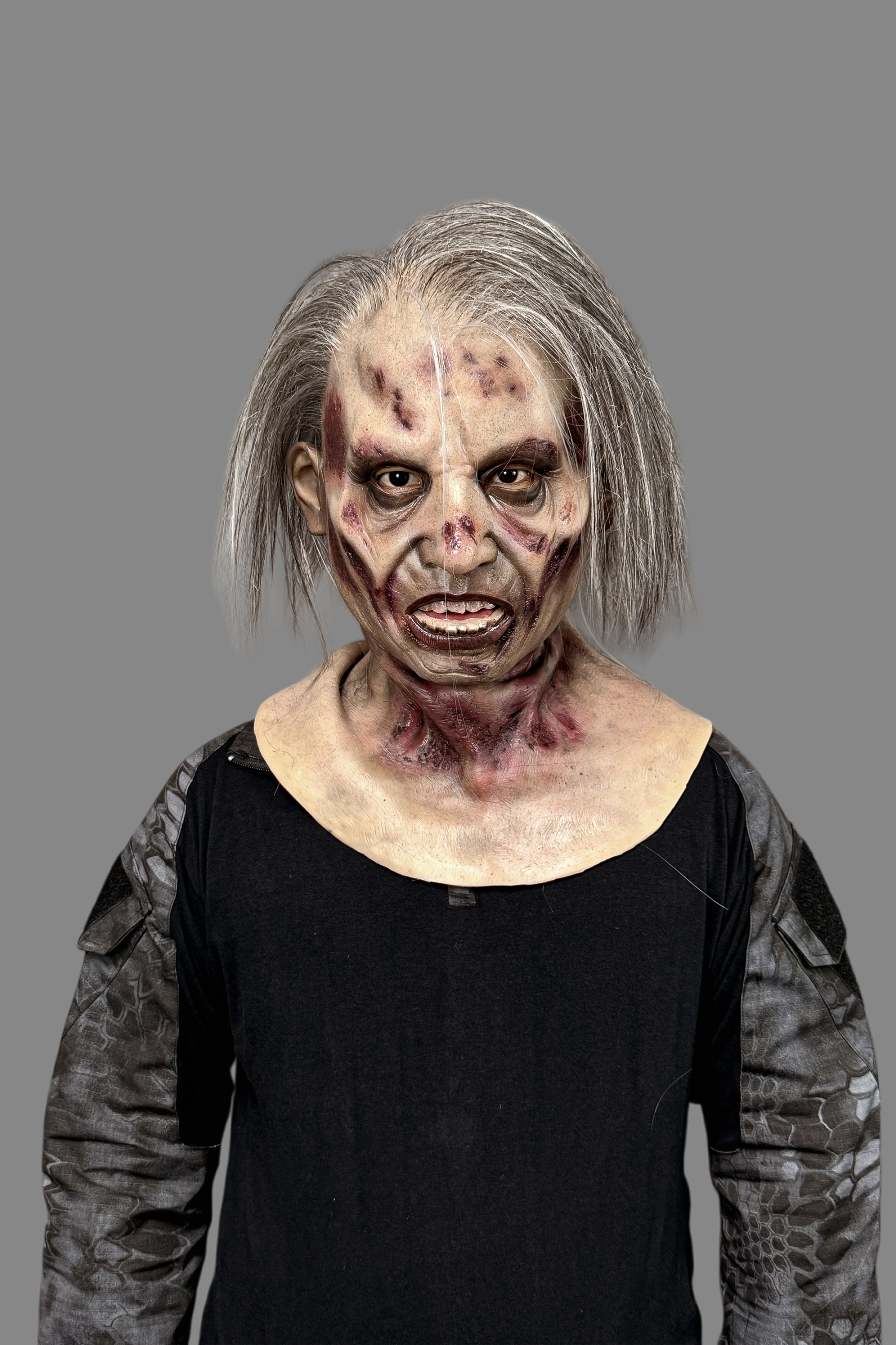 Hyper-realistic zombie old man silicone mask with long gray hair, rotting flesh, blood wounds and exposed teeth, worn with tactical black shirt.