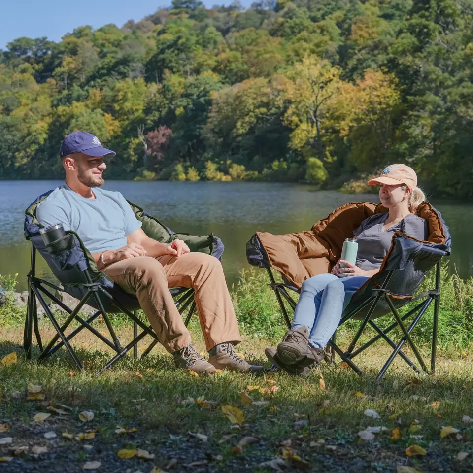 Man and a woman each relaxing in chairs by a lake.