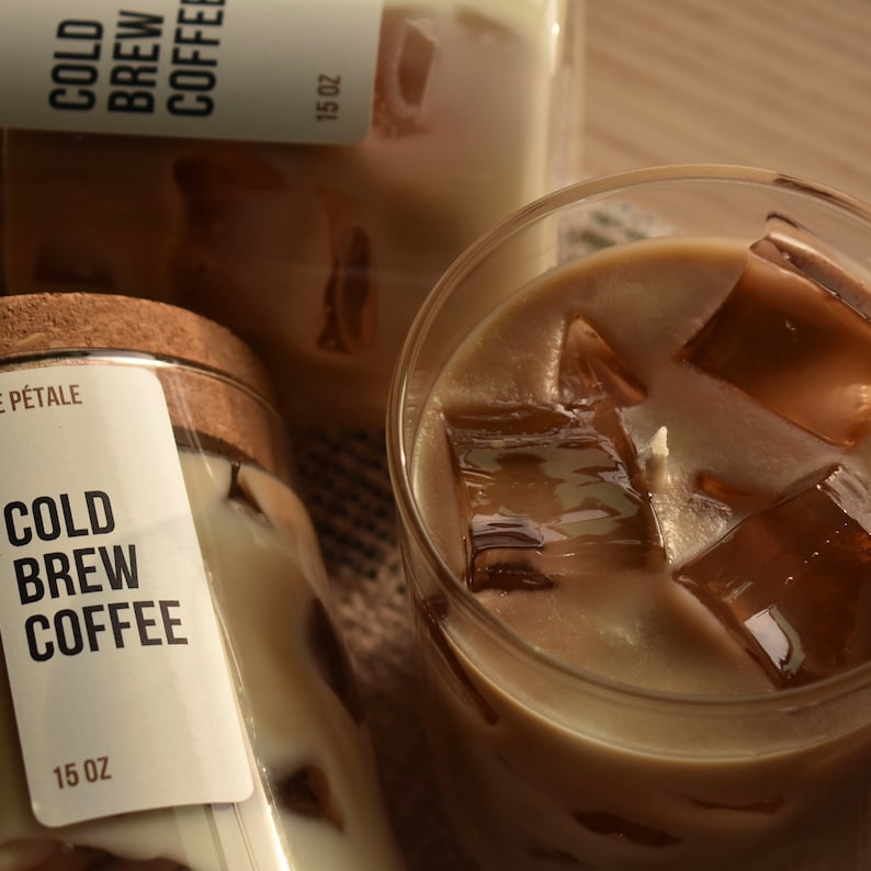 May include: Two glass jars with cork lids filled with a brown and white candle. The jars have labels that read 'Cold Brew Coffee' and '15 oz'. A glass cup filled with a brown liquid and ice cubes is in the foreground.
