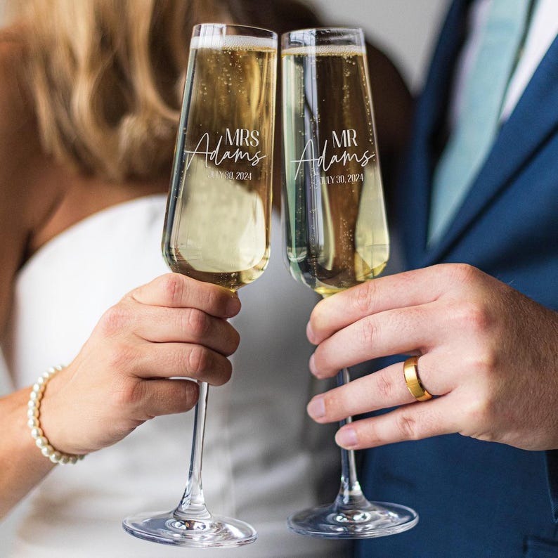 May include: Two champagne flutes with the words 'Mrs. Adams' and 'Mr. Adams' etched on them. The flutes are filled with champagne and are being held by a couple.