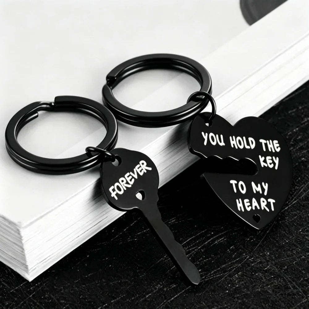 Two black metal keychains shaped like a broken heart and key, with white engraved text reading 'FOREVER' and 'YOU HOLD THE KEY TO MY HEART', displayed on stacked white books over a dark surface.