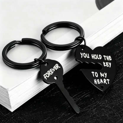 Two black metal keychains shaped like a broken heart and key, with white engraved text reading 'FOREVER' and 'YOU HOLD THE KEY TO MY HEART', displayed on stacked white books over a dark surface.