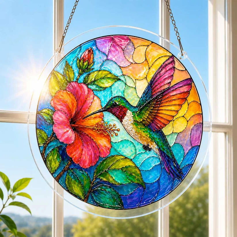 A round acrylic suncatcher with a hummingbird feeding from a large hibiscus flower hung in front of a sunny window