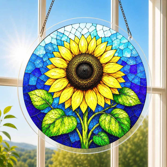 A round acrylic suncatcher with a large golden sunflower and green leaves hanging from a chain in front of a bright white window