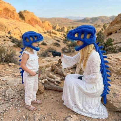 A woman and a child wearing matching blue dinosaur costume headsets and spiky back accessories pose outdoors in a rocky desert landscape. The woman wears a long white dress and black gloves, while the child wears a white sleeveless outfit.