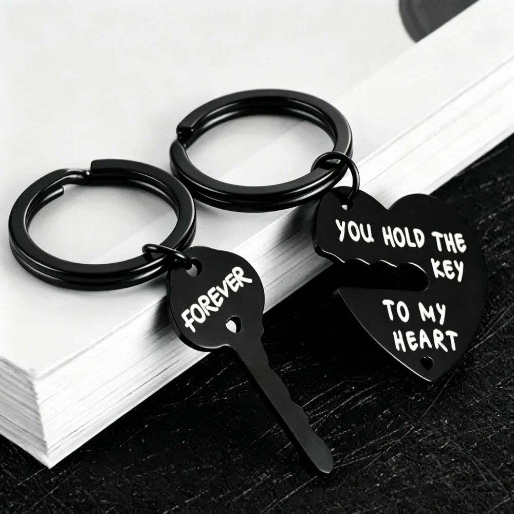 Two black metal keychains shaped like a broken heart and key, with white engraved text reading 'FOREVER' and 'YOU HOLD THE KEY TO MY HEART', displayed on stacked white books over a dark surface.