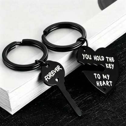 Two black metal keychains shaped like a broken heart and key, with white engraved text reading 'FOREVER' and 'YOU HOLD THE KEY TO MY HEART', displayed on stacked white books over a dark surface.
