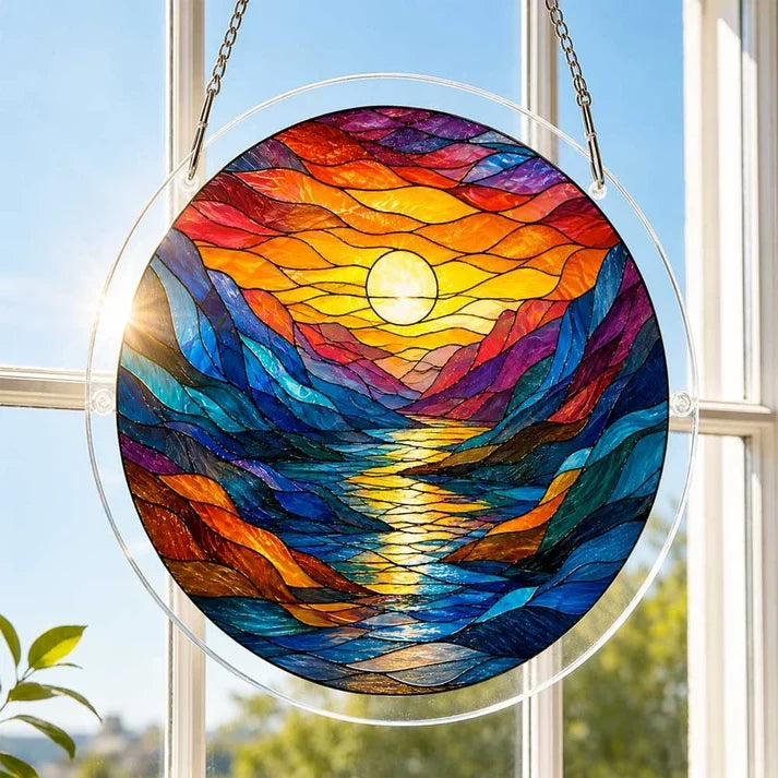 A round acrylic suncatcher with a colorful mountain river sunset and sun reflected on water hanging from a chain in front of a bright white window