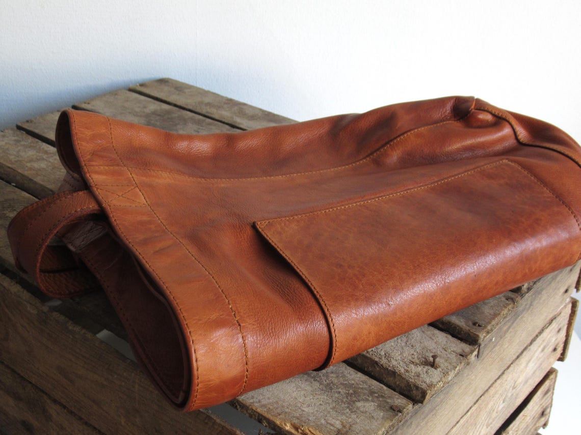 May include: A rich, brown leather tote bag rests on a rustic wooden crate. The bag features a spacious main compartment and a smaller exterior pocket. Its supple, full-grain leather shows subtle variations in tone, suggesting a handcrafted quality. The bag's simple design and sturdy construction highlight its timeless appeal. This versatile leather bag is perfect for everyday use or travel.