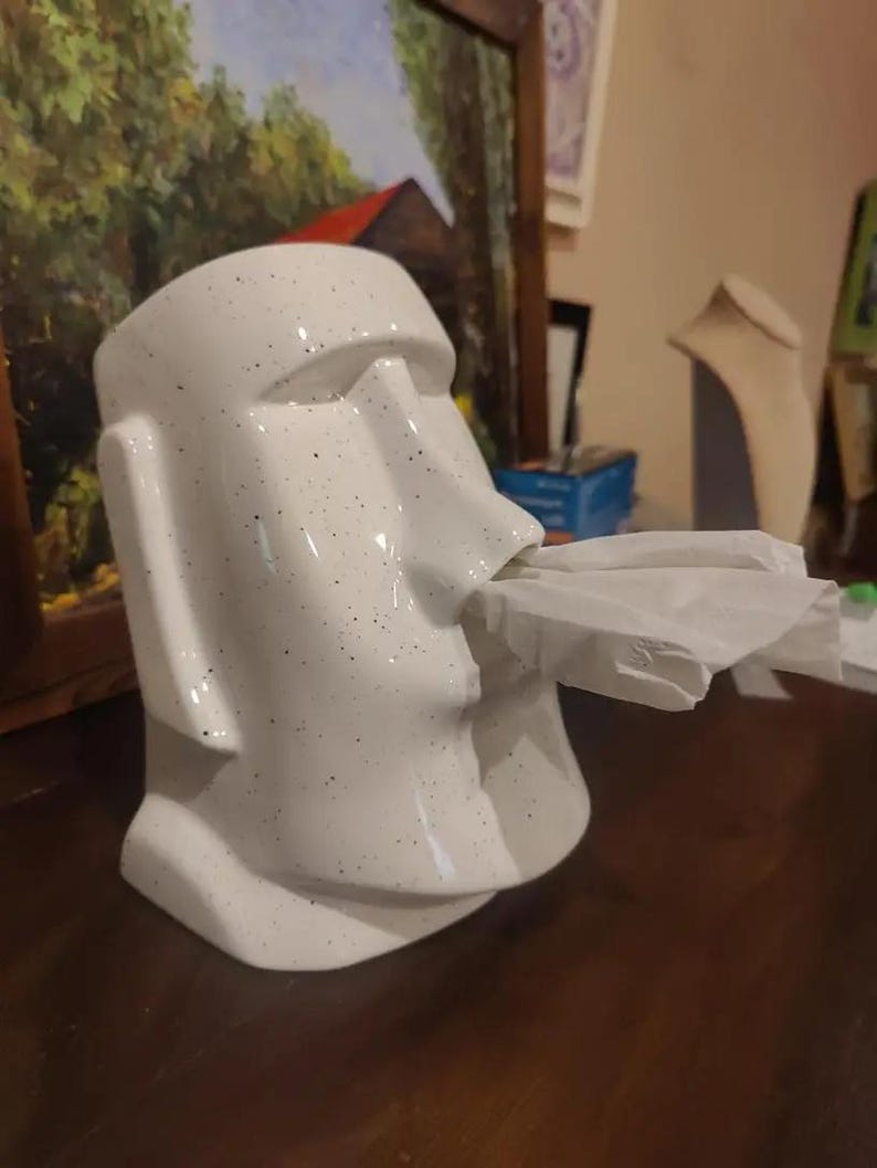 May include: White ceramic tissue box holder shaped like a Moai statue. The holder has a speckled finish and features a facial expression with a tissue-dispensing opening at the mouth. The tissue is partially visible, extending from the mouth of the statue. The holder is displayed on a dark brown surface.