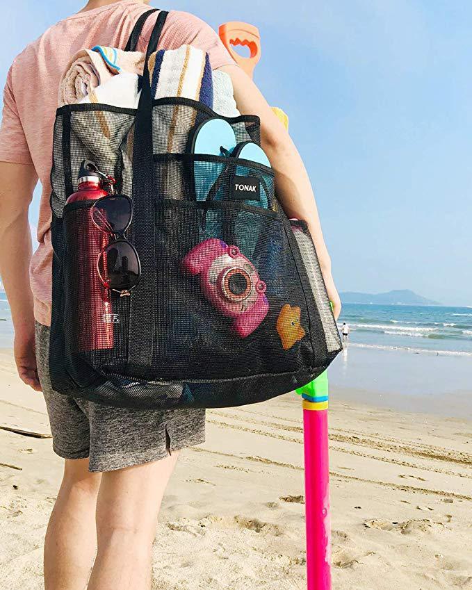 A man walking on a sandy beach carrying a sandproof mesh beach bag loaded with towels, sunglasses, and vacation gear.