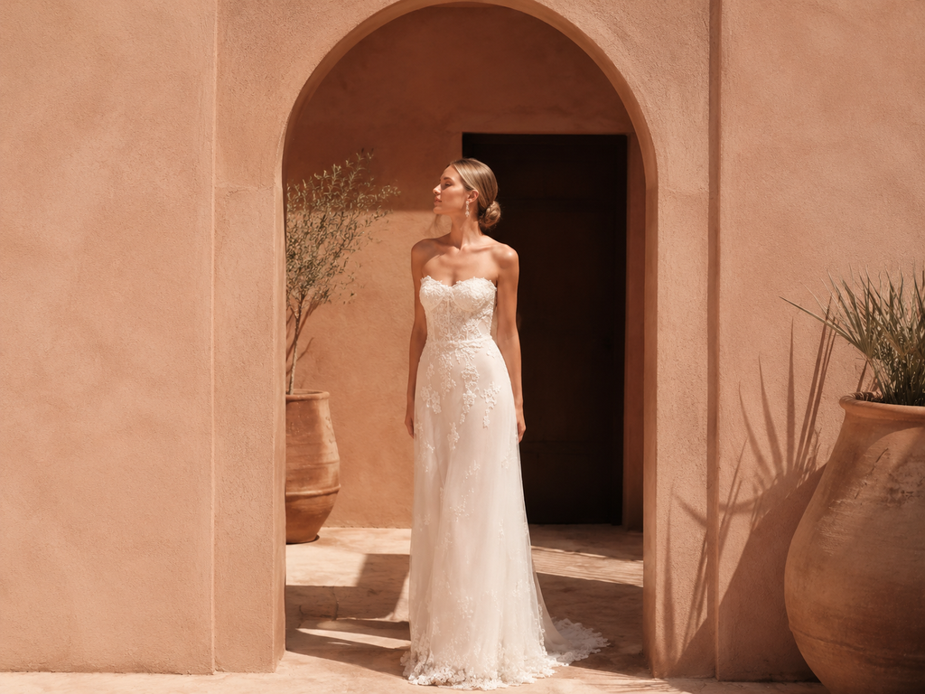 Bride wearing a simple strapless sheath wedding dress with delicate lace details, standing in a warm Mediterranean-style courtyard.