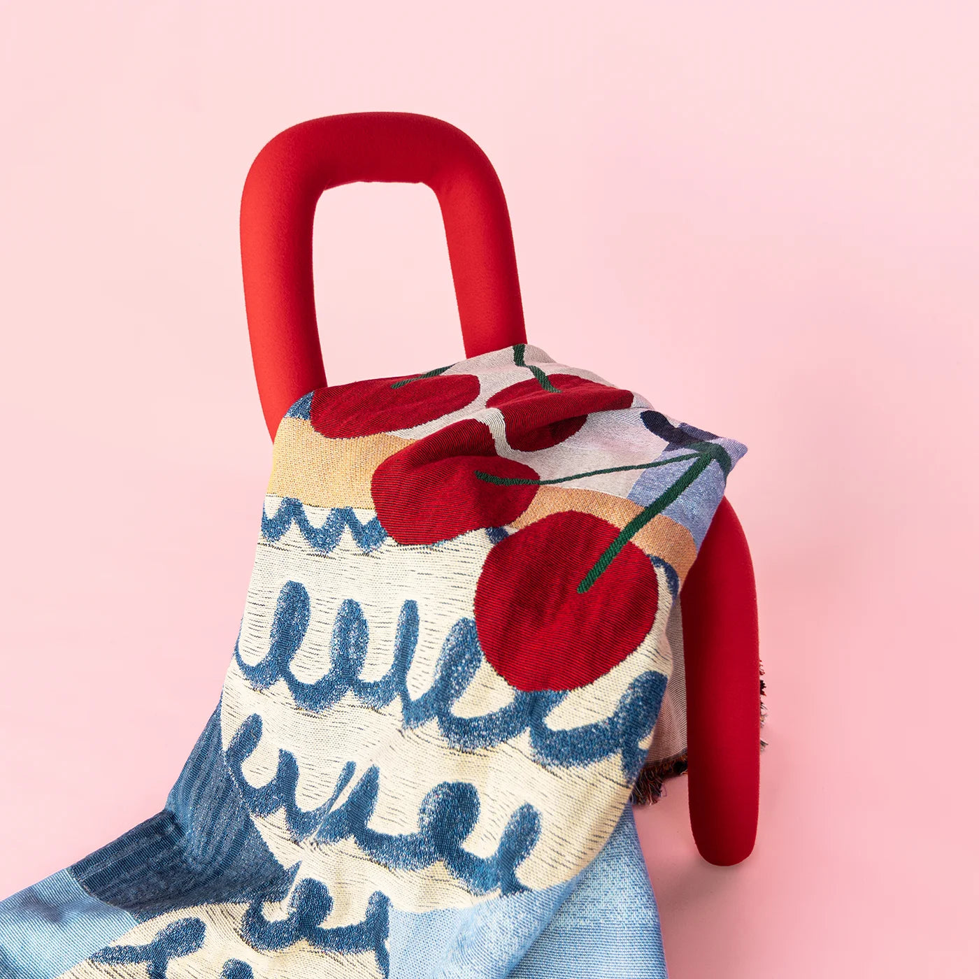 Artist-designed woven throw blanket with a blue vase & cherry design, draped over a red chair on a pink background.