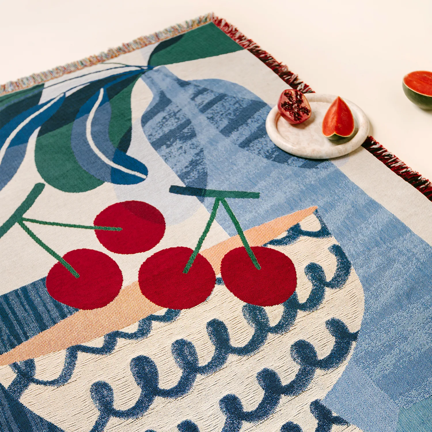 Flatlay of Slowdown Studio's artist-designed woven blanket, the Freya throw, featuring a blue, abstract fruit bowl with red cherries.
