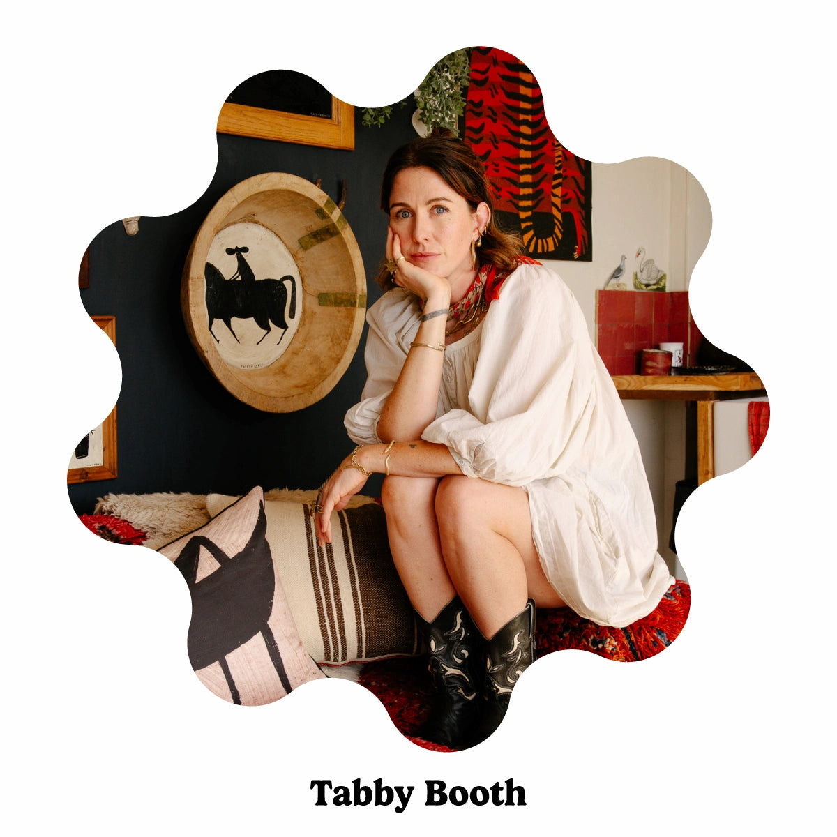 Female folk artist Tabby Booth in front of her artworks with black cowboy boots in a black and red living room.