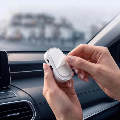 A person's hands holding a small white oval car accessory with a peeling cover in front of a car dashboard and windshield.