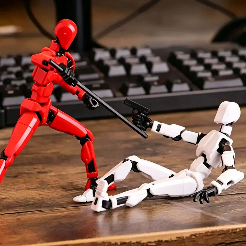 Two small humanoid action figures posed on a wooden tabletop with a blurred keyboard in the background. One figure is red and standing, holding a black gun.