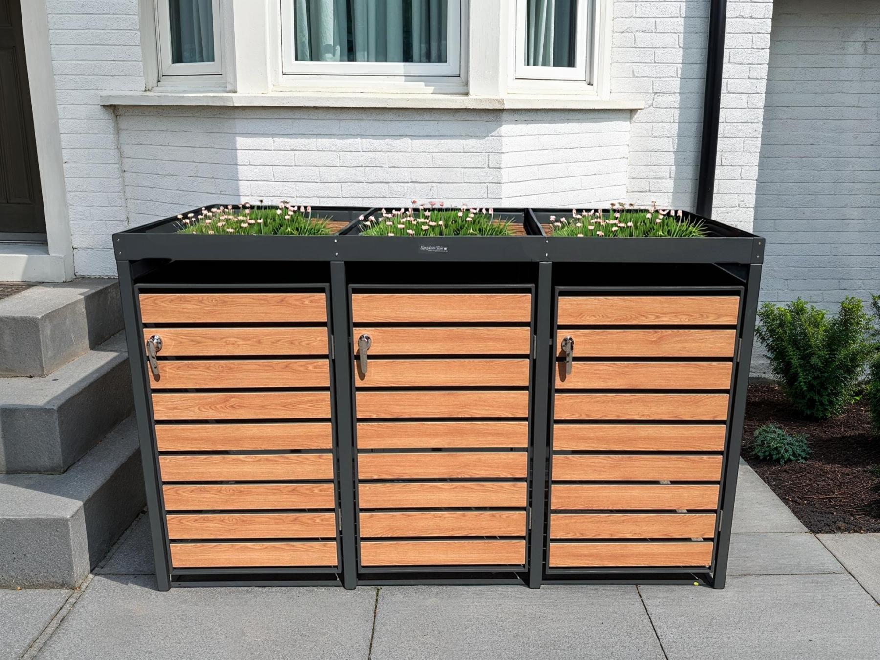 Triple wheelie bin store with wood-effect doors and integrated planter top positioned outside a white brick house.