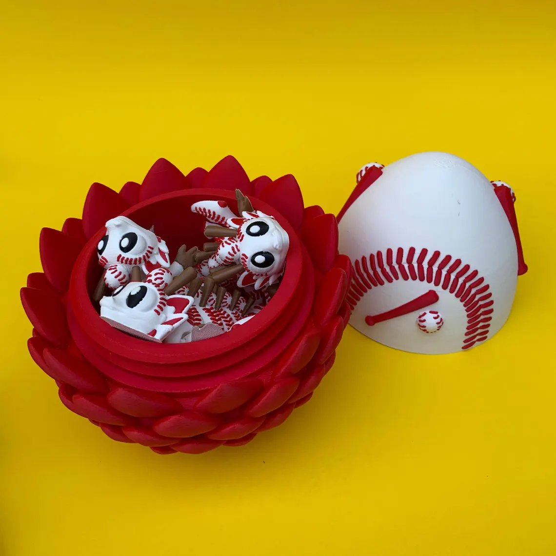 May include: A red, flower-shaped container holds several white and red baseball-themed figurines. A baseball-shaped container is open next to the flower. The background is a solid yellow.