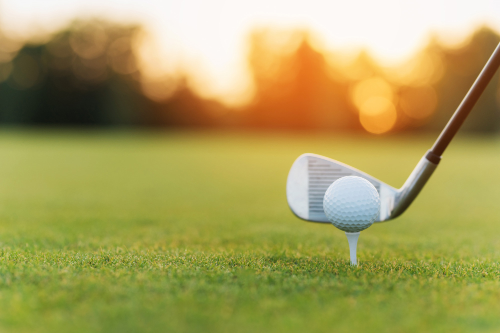 Golf ball on a tee with an iron behind it, sunset course in the background
