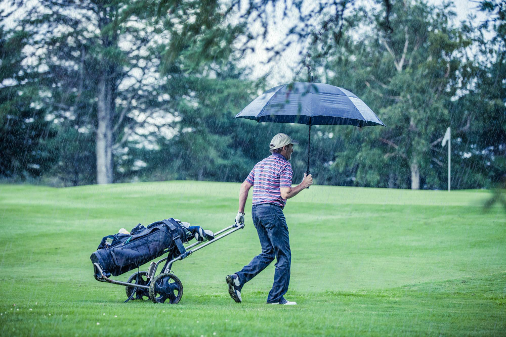Golfer walking in the rain with an umbrella while pulling a golf bag cart on the course