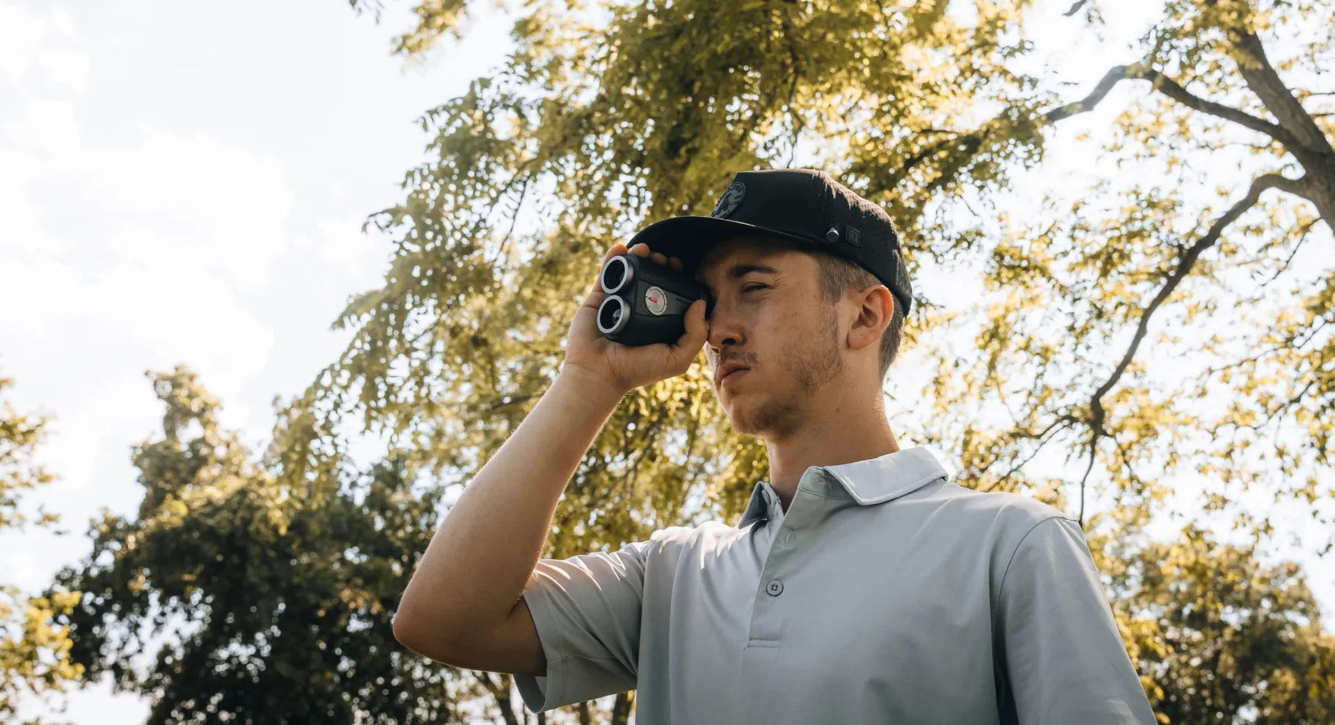 A golfer taking distance readings with a laser rangefinder outdoors under leafy trees