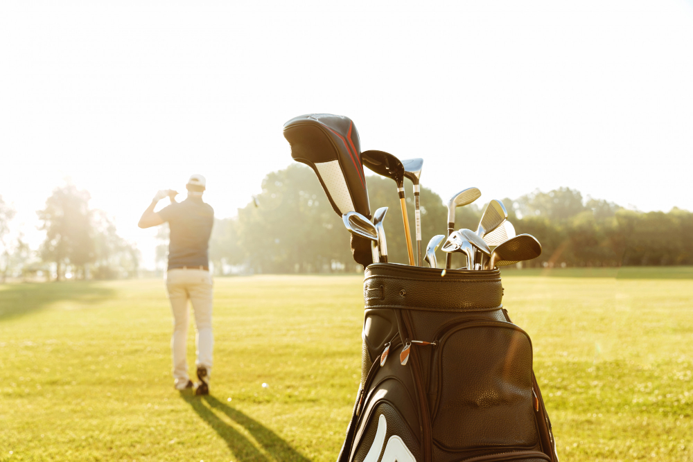 Golf bag with clubs in the foreground and a golfer mid-swing in the background on a sunlit course
